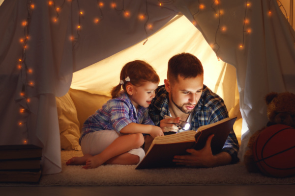 happy father and child daughter reading a book with a flashlight in a tent at home