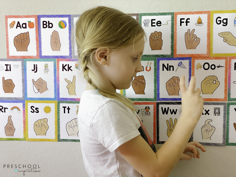 young girl trying to make the ASL alphabet letter n in front of colorful alphabet wall cards